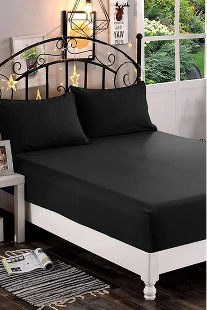 Ein ordentlich gemachtes Bett mit schwarzen Laken und Kissen, mit einem schwarzen, verzierten Metallkopfteil mit Sternverzierungen, neben einem Fenster mit Blick auf den Garten und einem Nachttisch mit einer Lampe und Büchern.