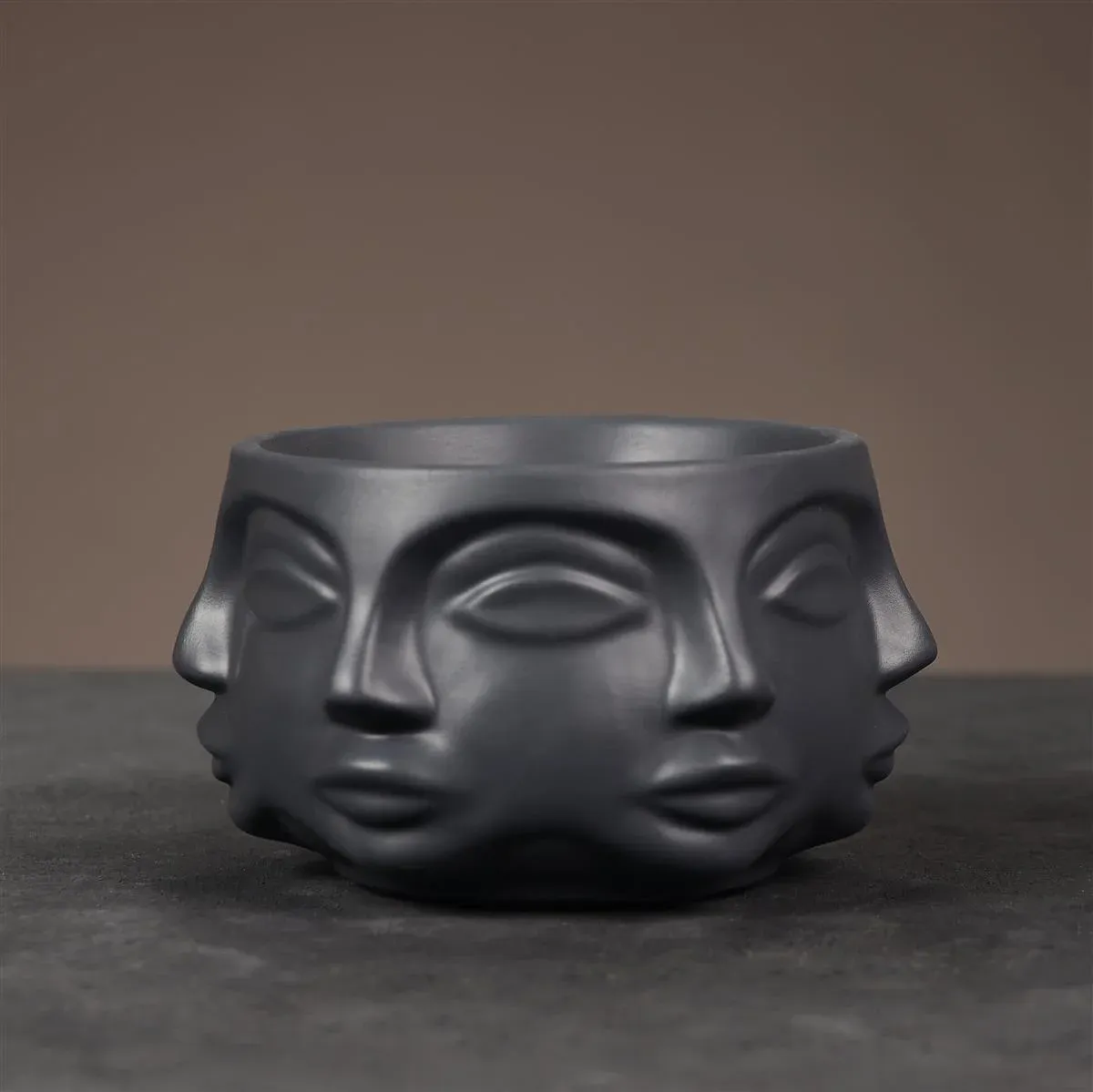 Eine dunkelgraue Keramikschüssel mit skulptierten Gesichtern, die auf einem braunen Hintergrund angeordnet sind.