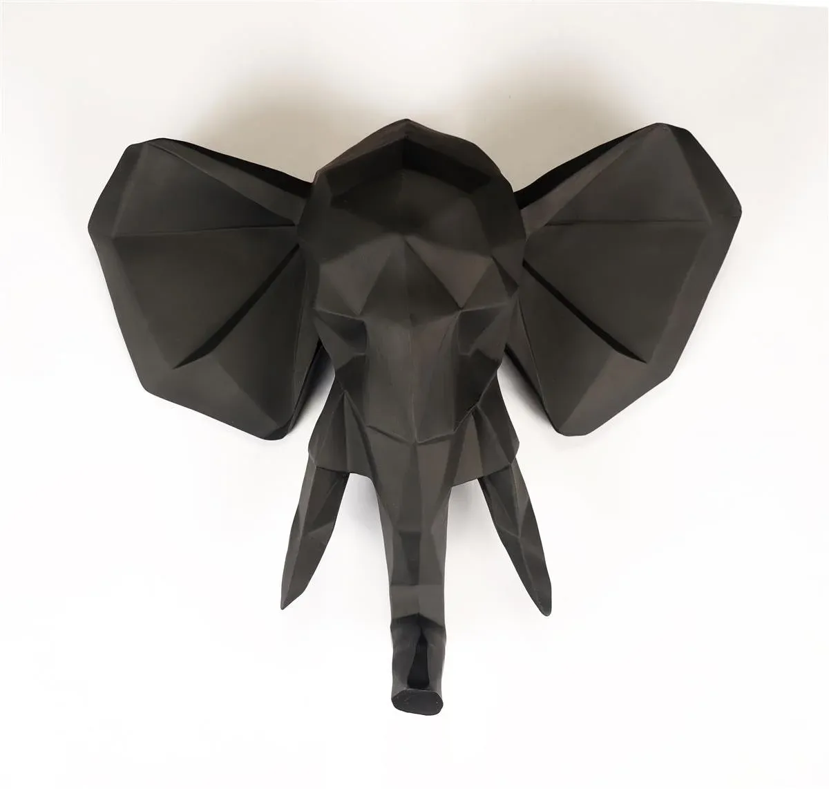 Eine geometrische, schwarze Elefantenkopfskulptur im Origami-Stil ist vor einem weißen Hintergrund angebracht. Das Design weist kantige Facetten und stilisierte Formen für Ohren, Rüssel und Stoßzähne auf.